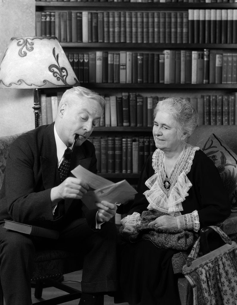 Detail of 1930s 1940s retired senior couple looking at check in living room man smoking pipe by Anonymous