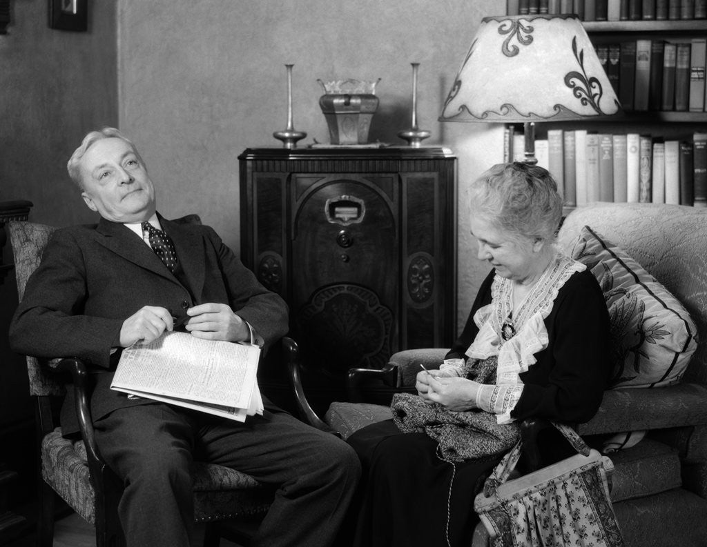 Detail of 1930s elderly couple sitting in living room listening to radio woman knitting man with newspaper in lap by Anonymous
