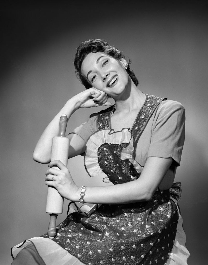Detail of 1950s woman housewife wearing apron holding rolling pin looking at camera by Anonymous