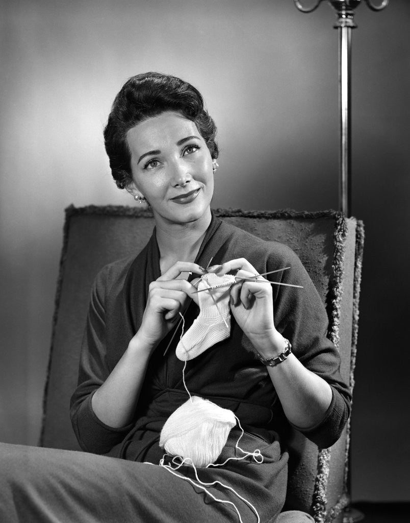 Detail of 1950s woman crocheting baby sock looking at camera by Anonymous