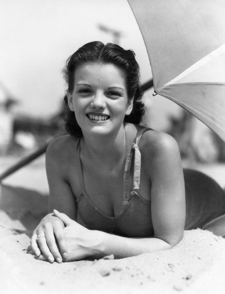 Detail of 1930s teen girl lying on beach under umbrella wearing bathing suit smiling looking at camera by Anonymous