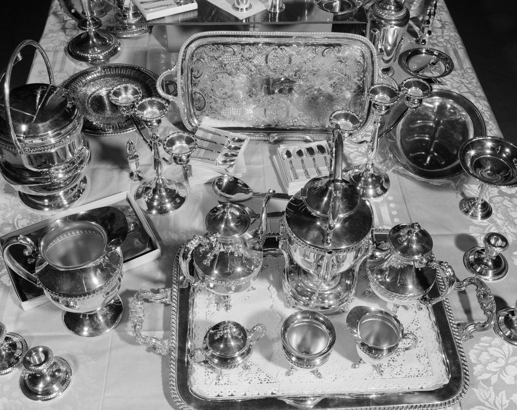 Detail of 1950s still life overhead view of assortment of silver service pieces including tea set candlesticks & ice bucket by Anonymous