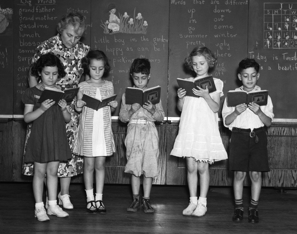 Detail of 1930s line-up of 5 elementary school students in front of blackboard reading books with teacher looking on by Corbis