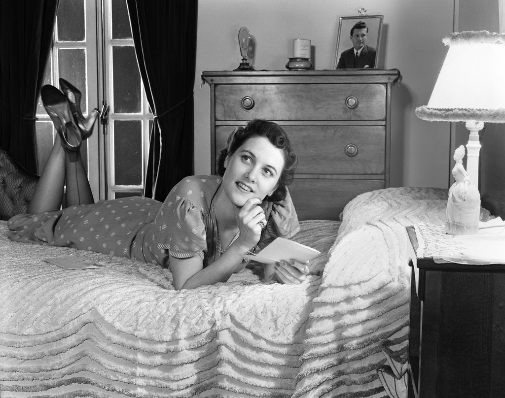 Detail of 1940s fully dressed smiling woman laying on her bed with her legs up reading a letter in her bedroom by Anonymous