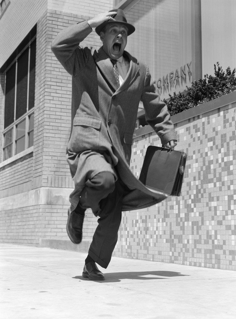 Detail of 1960s 1950s frantic man running down street holding hat on with hand carrying briefcase wearing top coat by Anonymous
