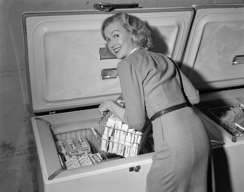 Detail of 1950s blond woman lifting wire basket food items from a deep freezer looking at camera over her shoulder by Anonymous