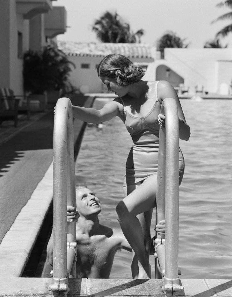 Detail of 1930s woman wearing one piece bathing suit climbing out of swimming pool looking down at man smiling by Anonymous