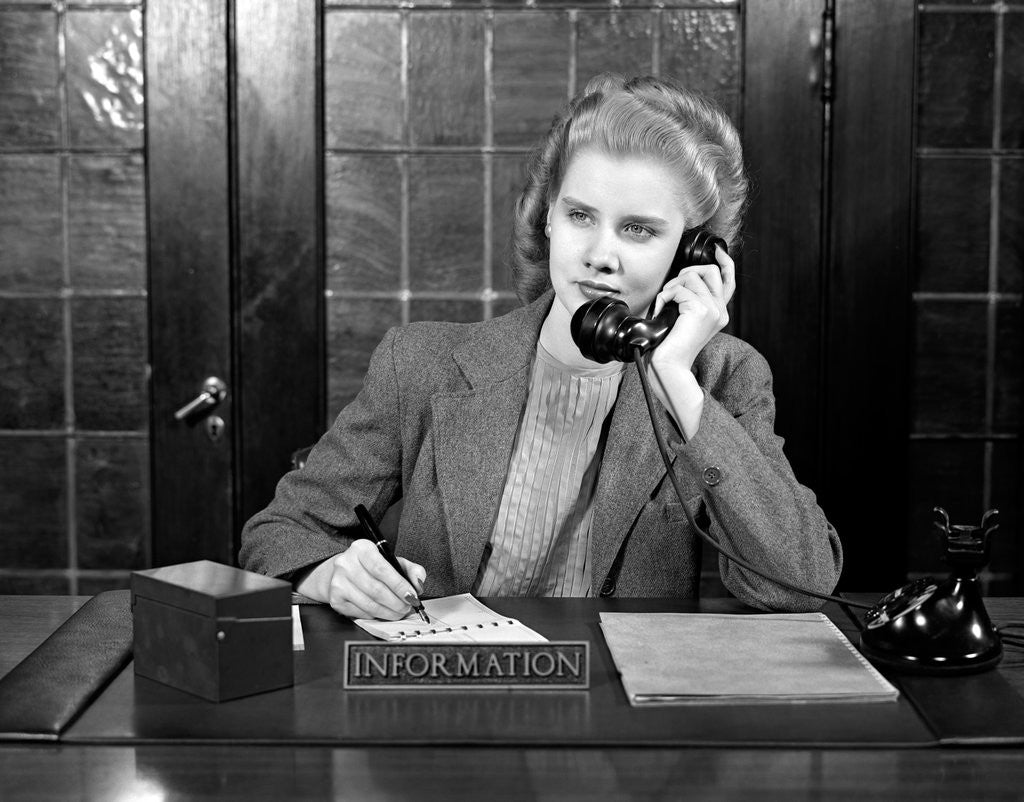 Detail of 1940s young woman sitting at information desk secretary talking on telephone writing by Corbis