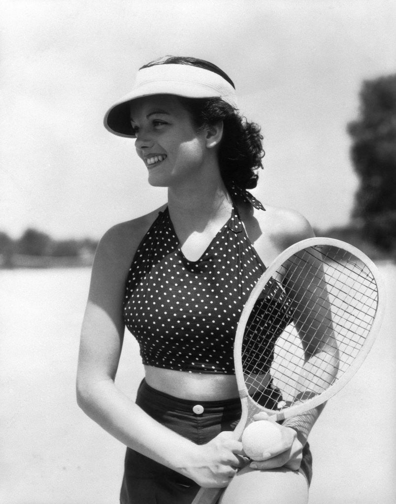 Detail of 1930s woman in polka dot halter top shorts & sun visor holding a tennis ball & racket by Anonymous