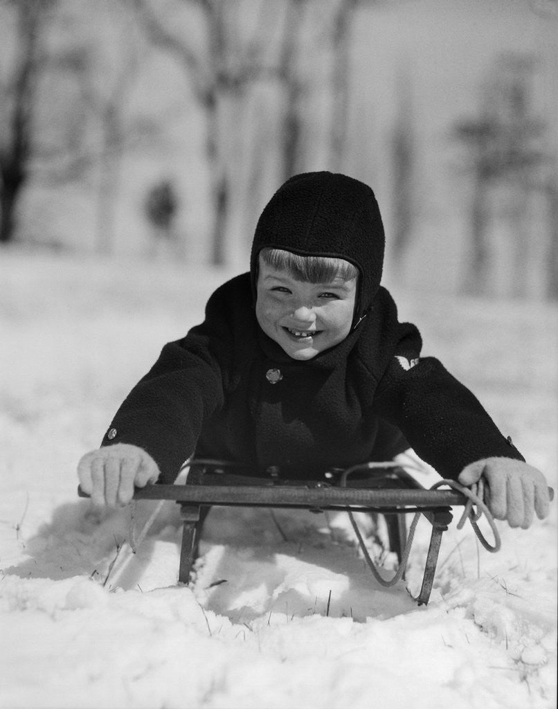 Detail of 1930s young smiling boy on sled in snow looking at camera by Anonymous