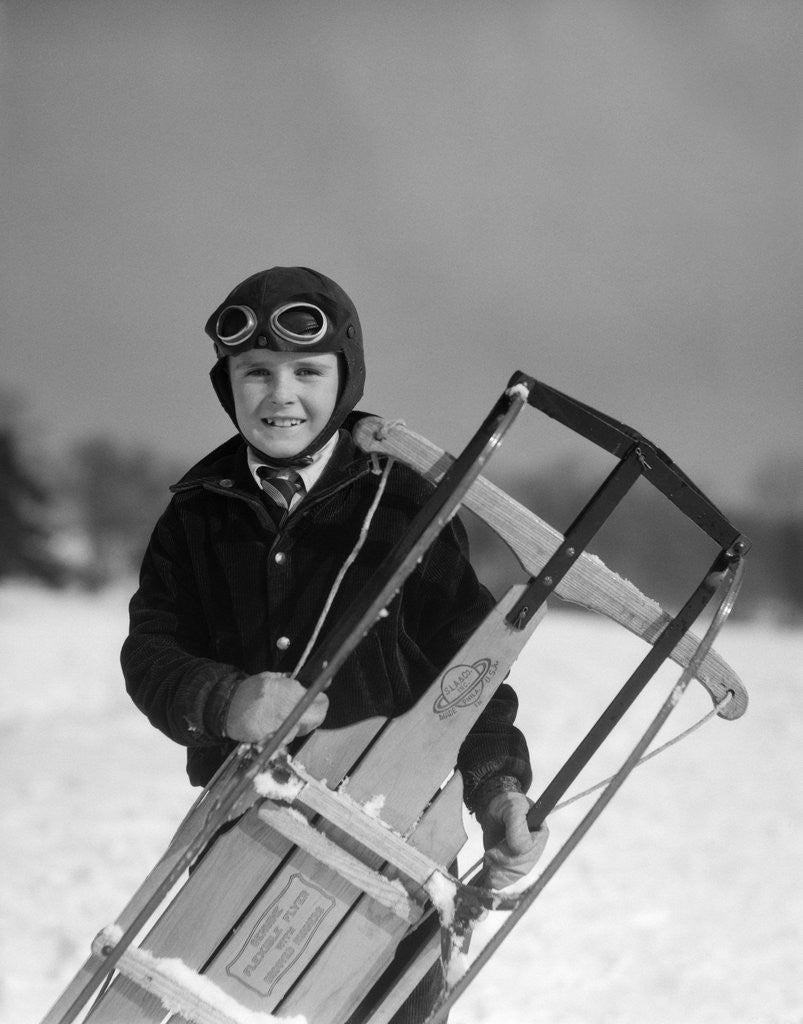 Detail of 1920s 1930s smiling boy wearing aviator goggles leather flying helmet holding sled standing in snow field looking at camera by Anonymous
