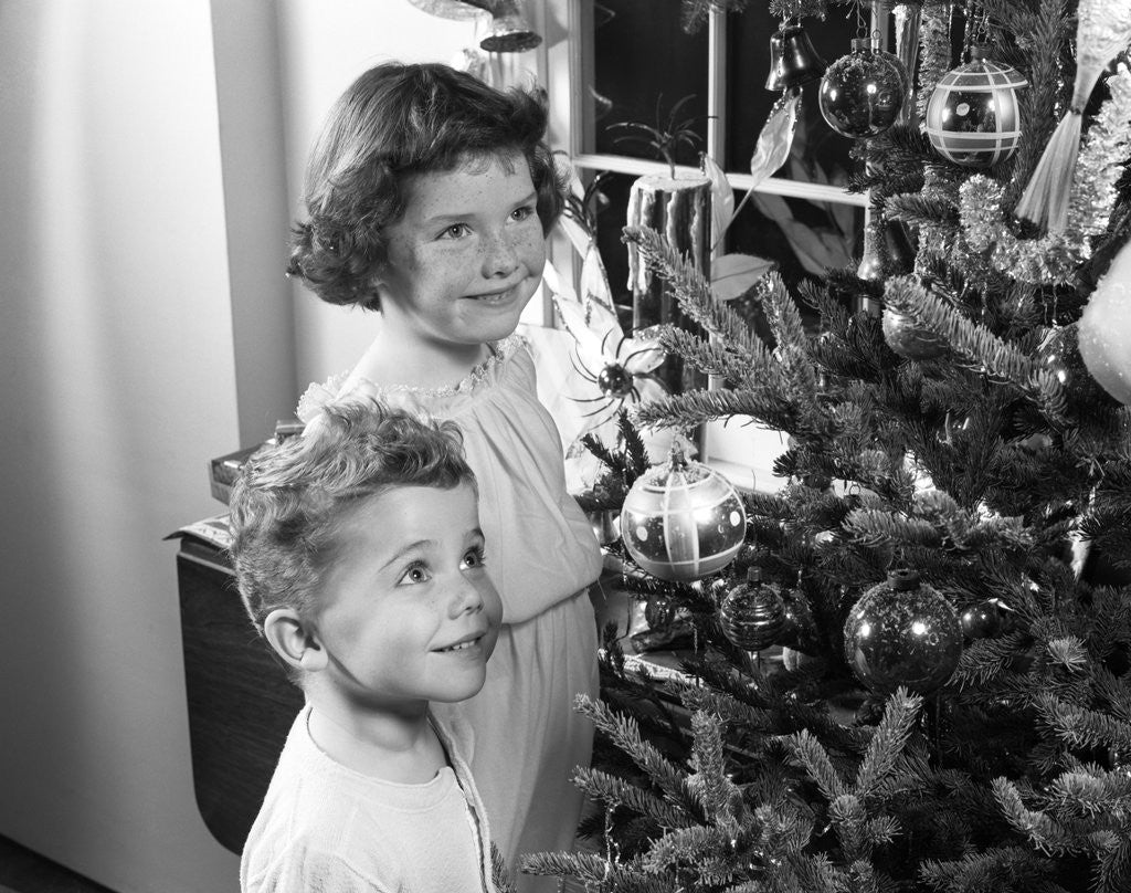 1950s boy girl wearing pajamas smiling up at christmas tree decorations by window by Corbis