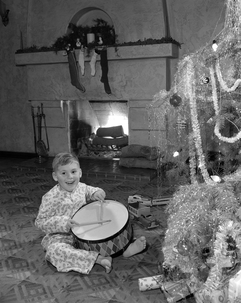 Detail of 1950s little boy playing toy drum by christmas tree and fireplace looking at camera by Anonymous
