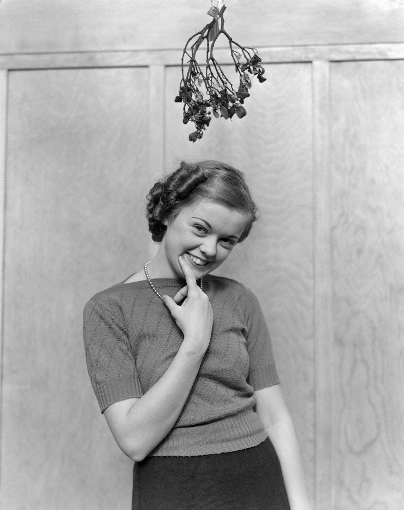 Detail of 1930s 1940s cute young woman standing under mistletoe smiling hand near mouth looking at camera by Corbis
