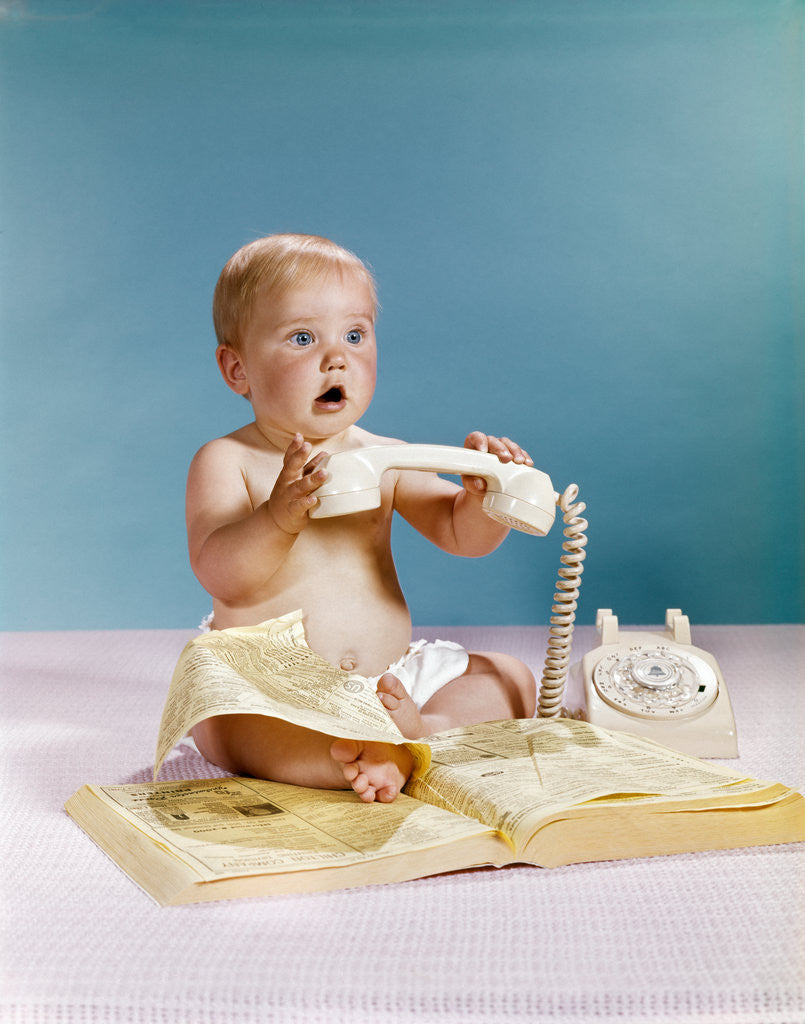 Detail of 1960s baby surprised facial expression holding telephone hand set over yellow pages phone book directory by Anonymous