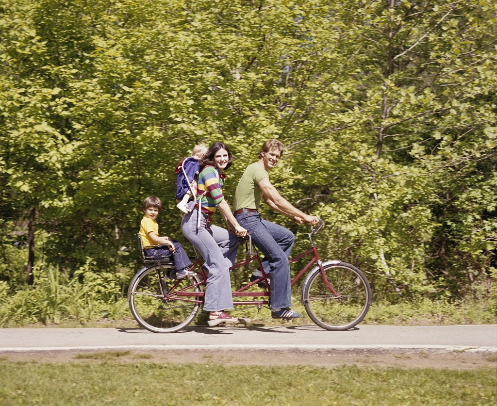 Detail of 1970s Family On Tandem Bicycle Mother Father Son & Baby Daughter In Backpack Wearing Bellbottom Blue Jeans Looking At Camera by Anonymous