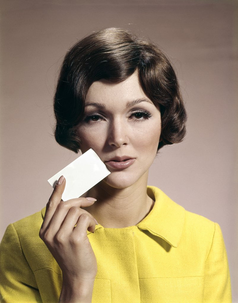 Detail of 1960s Brunette Woman Holding Blank Business Card To Face Looking At Camera Wearing Yellow Top by Anonymous