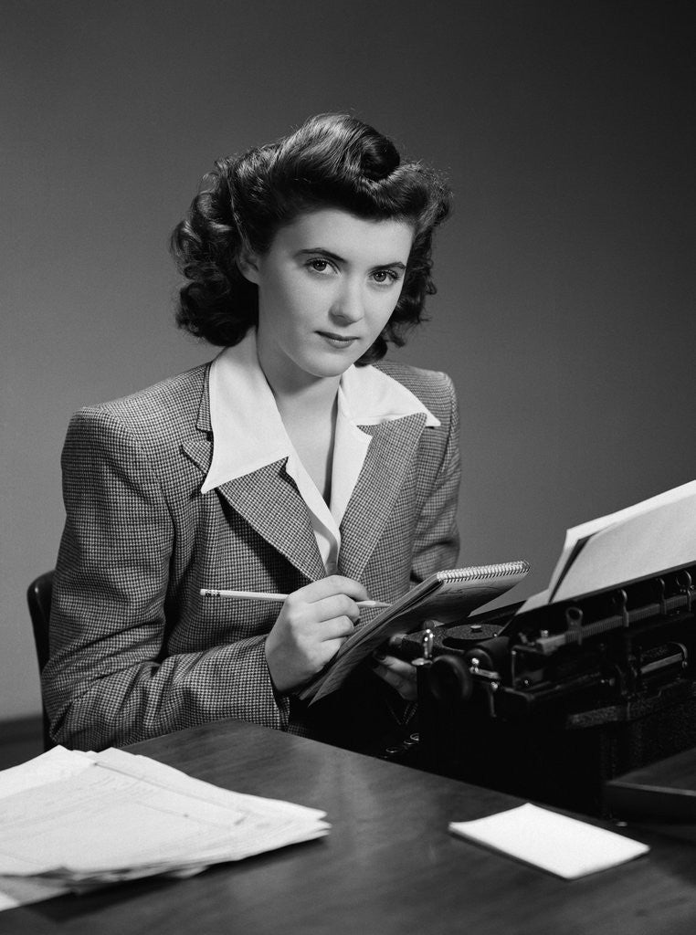 Detail of 1940s Serious Secretary Stenographer Woman With Pencil Checking Notepad Sitting At Desk With Manual Typewriter Looking At Camera by Corbis