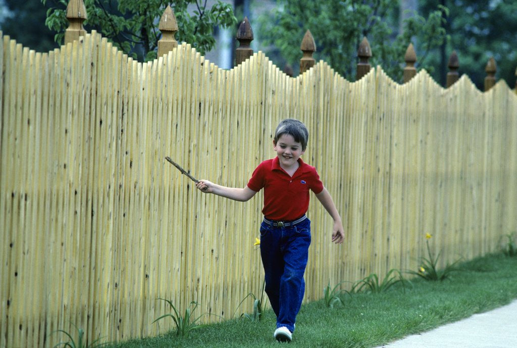 Detail of 1980s Smiling Boy Running Along Sidewalk Rattling Stick On Tall Picket Fence Making Noise by Corbis