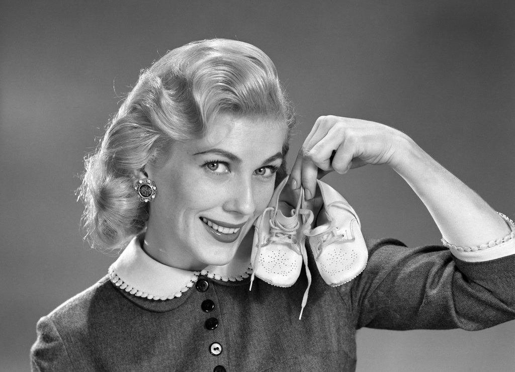 Detail of 1950s 1960s Smiling Blond Woman Communicating That She Is Mother To Be By Holding Up A Pair Of Baby Shoes Looking At Camera by Anonymous