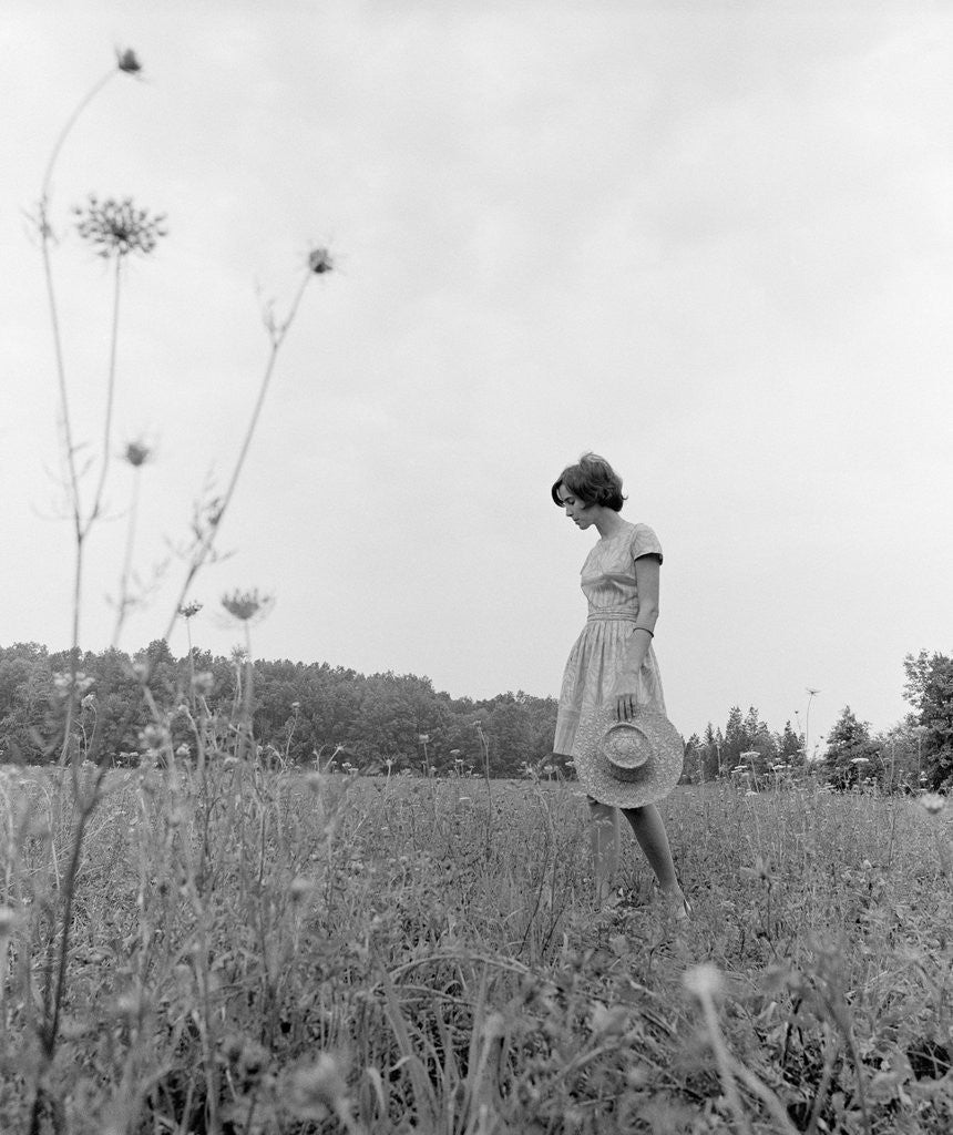 Detail of 1970s Woman Wearing Dress Carrying Large Straw Hat Standing In Middle Of Field Looking Down by Anonymous