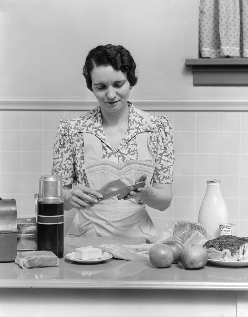 Detail of 1930s Woman Housewife In Kitchen Wearing Apron Making Sandwich Packing Metal Lunch Box Thermos Apples Bread Milk On Counter by Anonymous