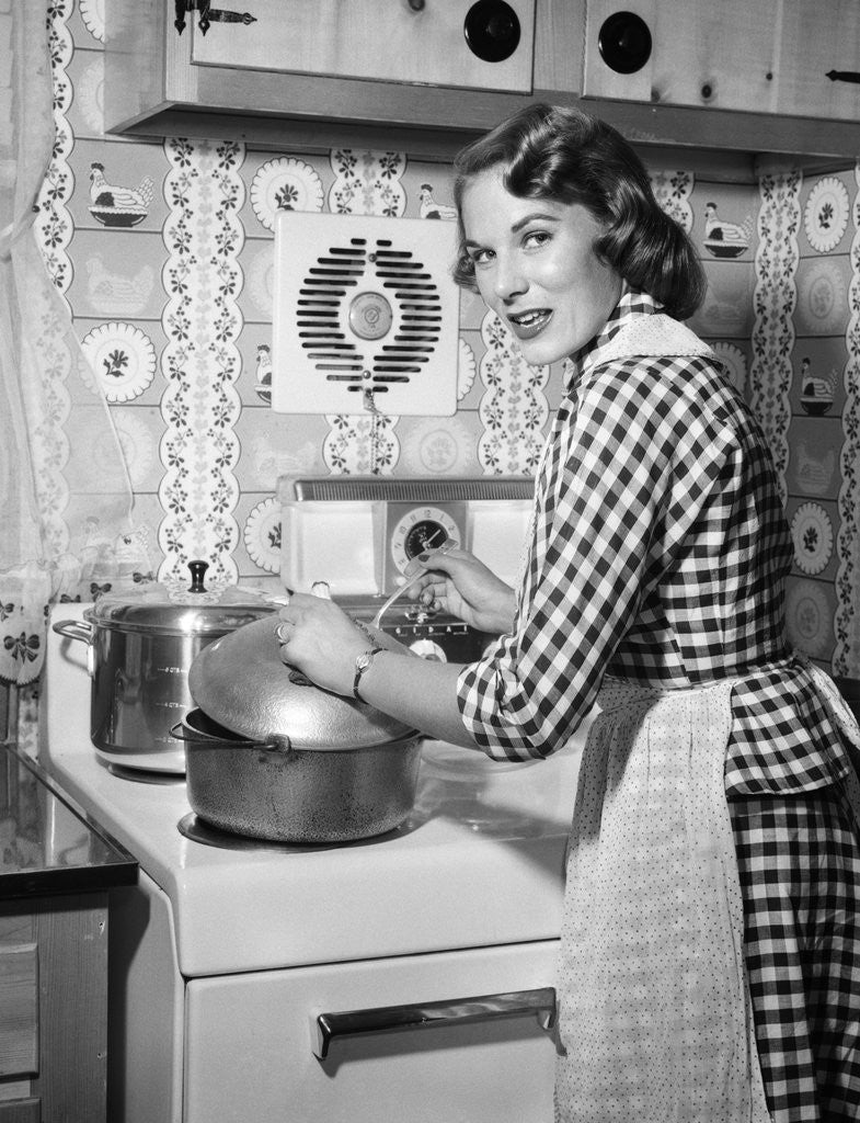 Detail of 1950s Housewife Wearing Checkered Dress Standing In Kitchen Stirring Pot On Stove Looking Over Shoulder by Anonymous