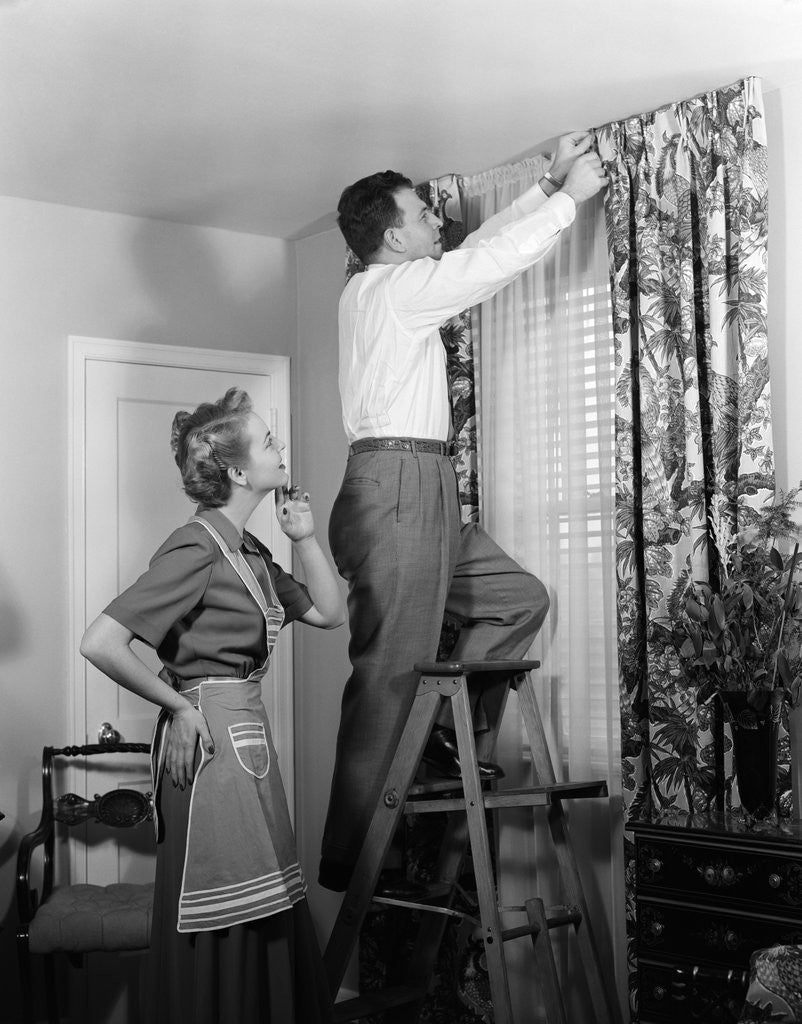 Detail of 1950s Skeptical Wife In Apron Fingers Pressing Chin Watching Husband On Ladder Hanging New Jungle Motif Drapes In Living Room by Anonymous