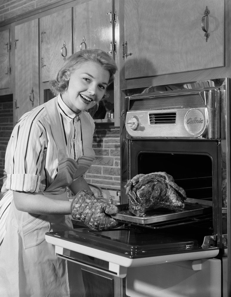 Detail of 1950s Smiling Woman Housewife Wearing Apron Taking Large Roast Beef From Electric Oven In Kitchen Looking At Camera by Anonymous