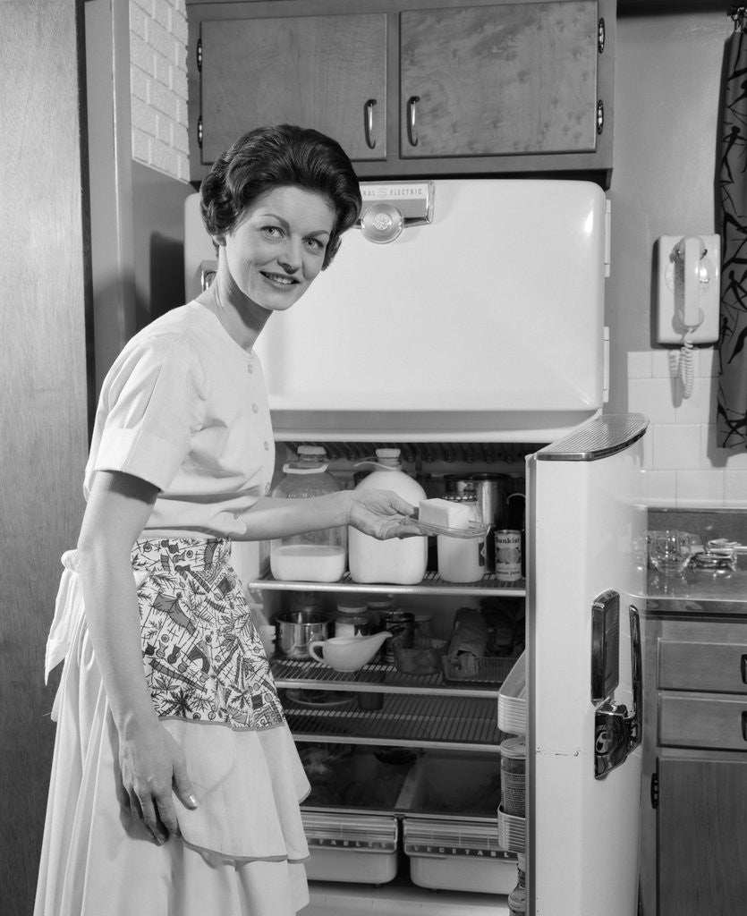 Detail of 1950s Smiling Woman Housewife Putting Stick Of Butter Into Electric Refrigerator In Kitchen Looking At Camera by Anonymous