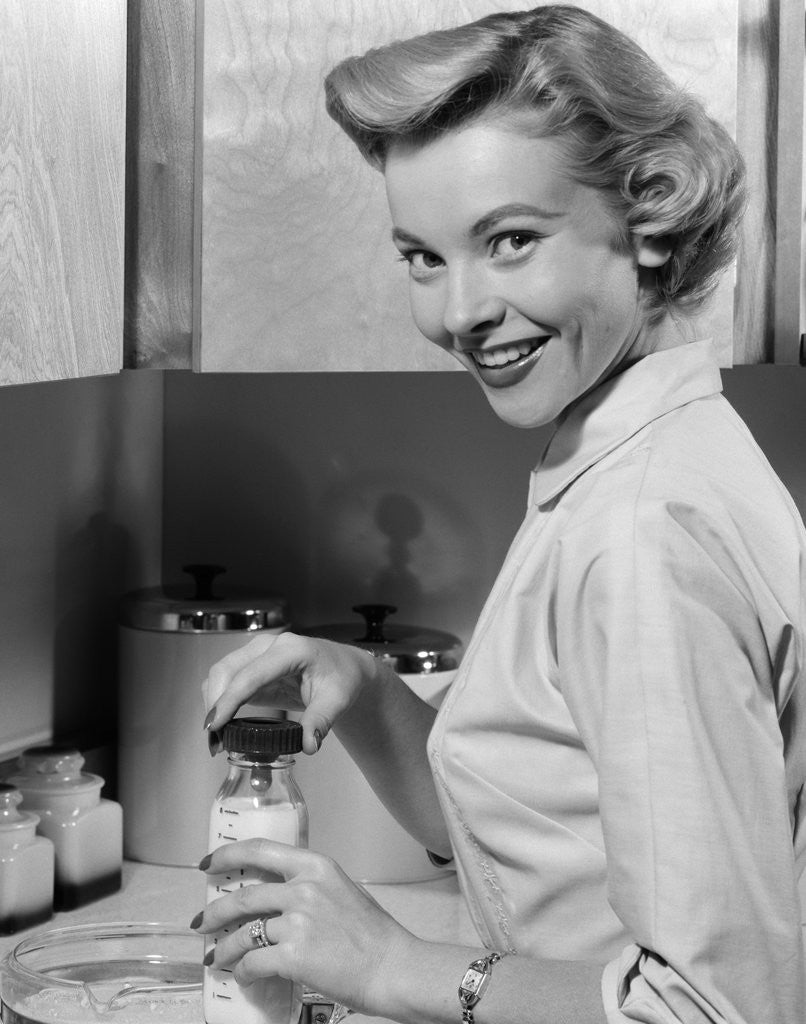 Detail of 1950s Smiling Blonds Woman Mother Warming A Baby Bottle Of Milk In Kitchen Looking At Camera by Anonymous