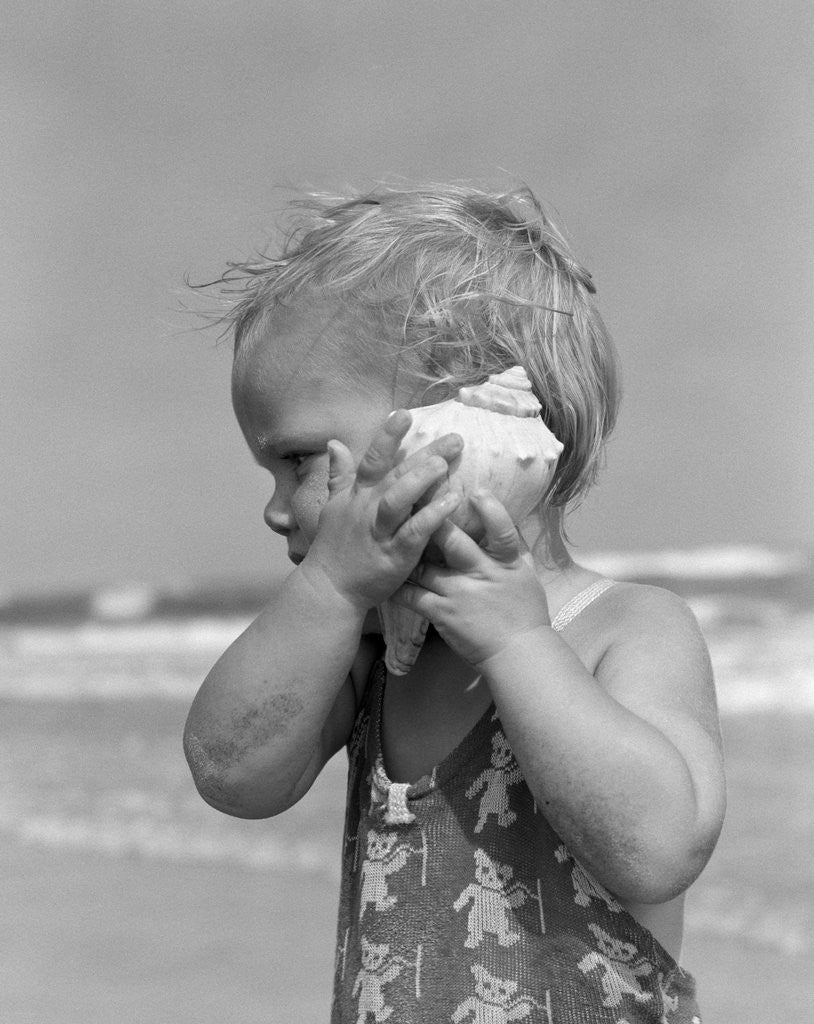 Detail of 1950s Blond Toddler Girl Listening To Ocean In A Seashell Wearing Teddy Bear Bathing Suit by Corbis