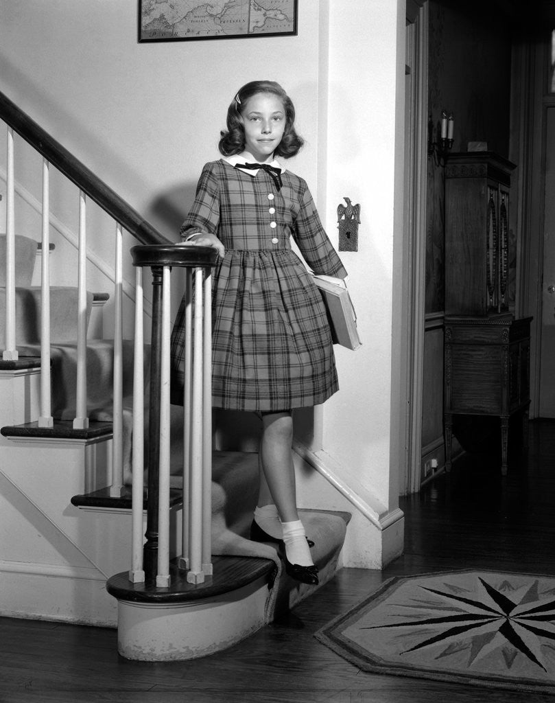 Detail of 1950s Teenage Girl In Plaid Dress And White Ankle Socks On Stairway Holding Banister Carrying School Books Looking At Camera by Anonymous