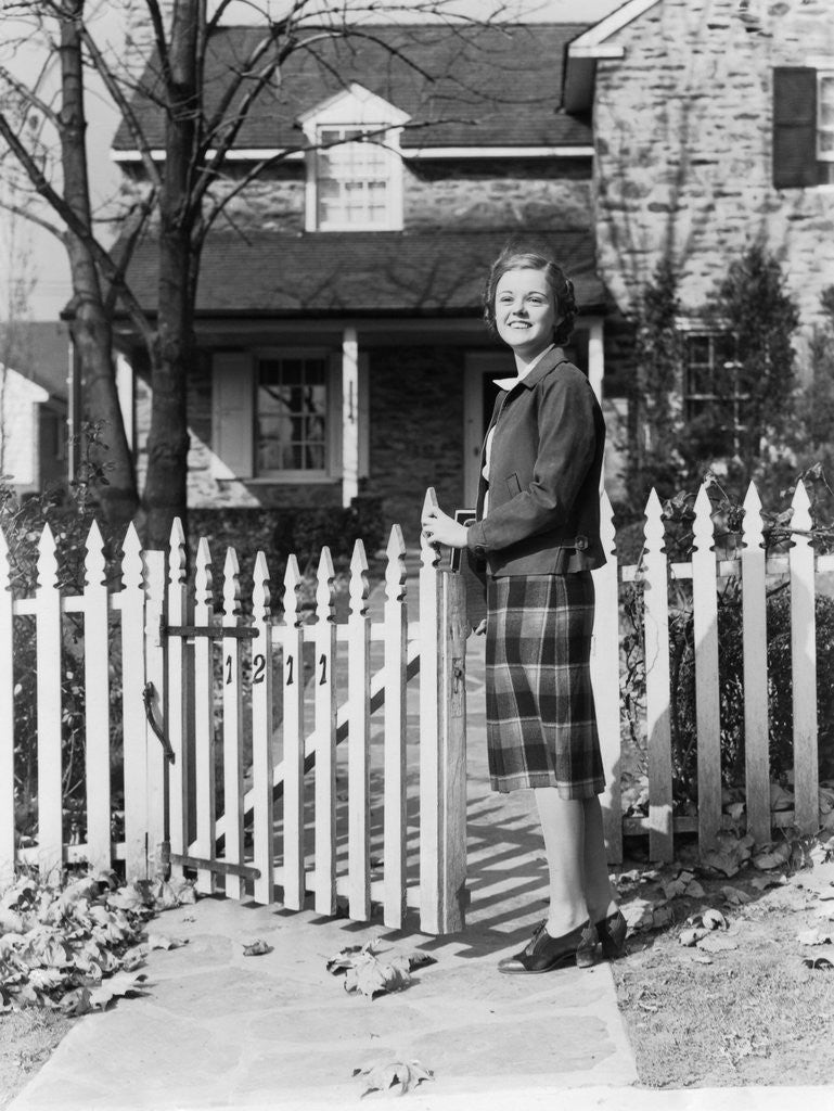 Detail of 1940s Smiling Pretty Young Teenage Girl Standing By White Picket Fence In Front Of Stone House In Autumn Looking At Camera by Anonymous
