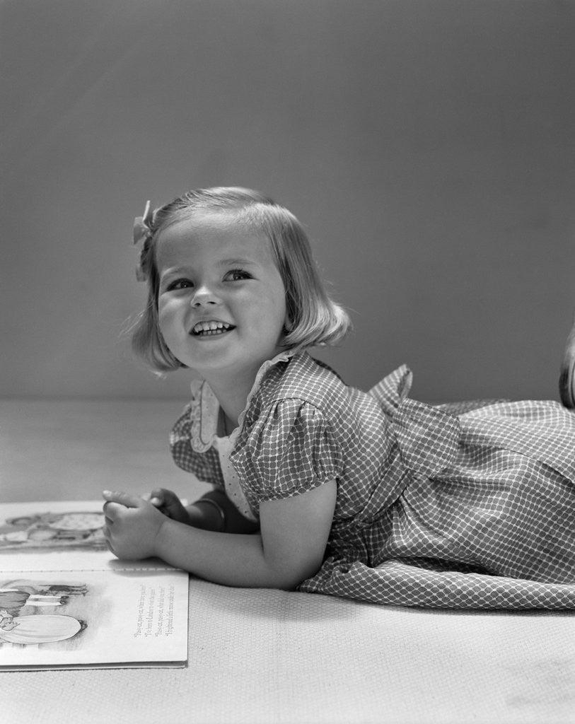 Detail of 1940s Little Blond Girl Lying On Floor Looking Up And Aside Smiling From Reading A Picture Book by Corbis