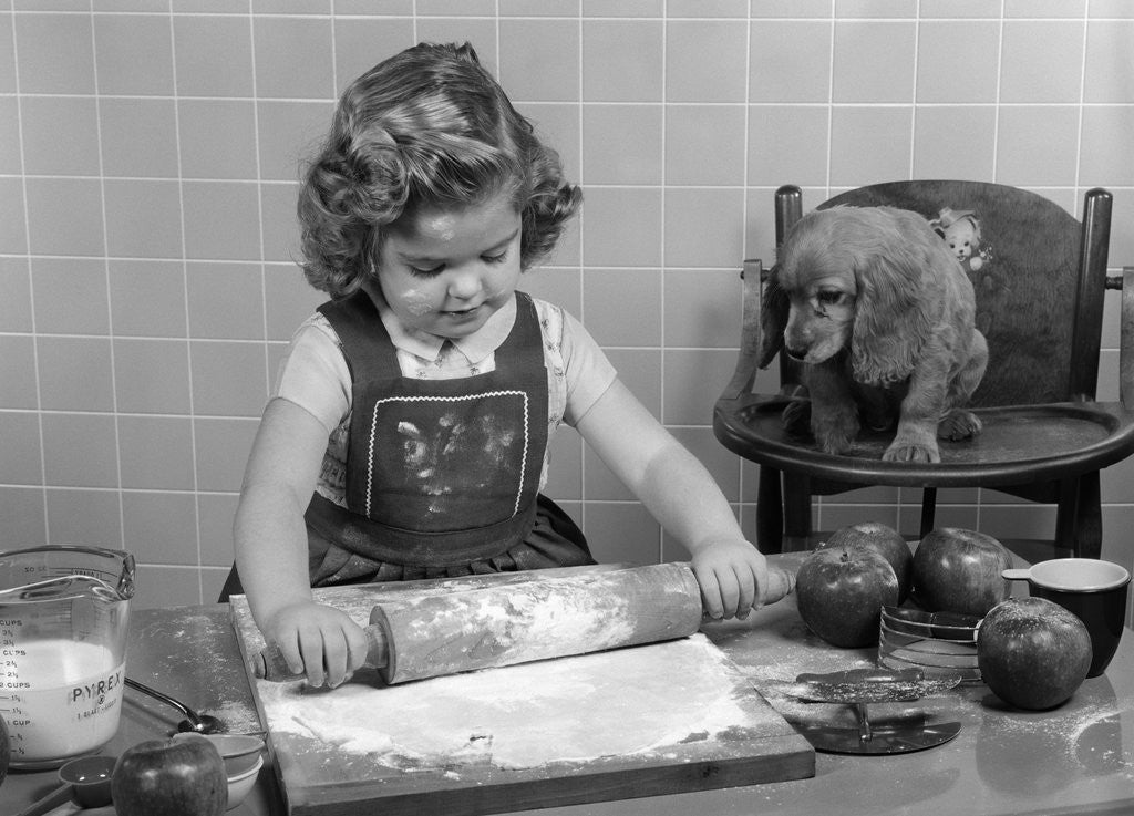 Detail of 1950s Little Girl Rolling Out Apple Pie Crust On Kitchen Table With Cocker Spaniel Puppy Watching by Anonymous