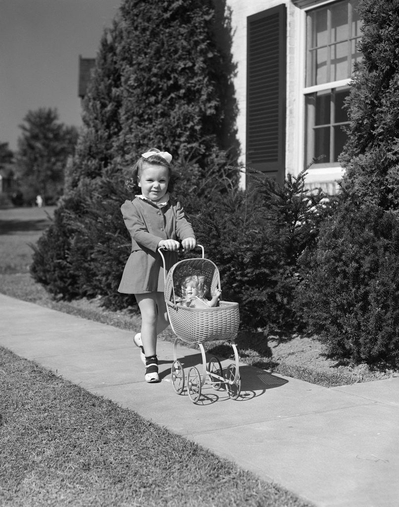 Detail of 1940s Little Girl Walking Pushing Her Doll In Antique Woven Wicker Stroller Looking At Camera On Sidewalk In Front Of House by Corbis