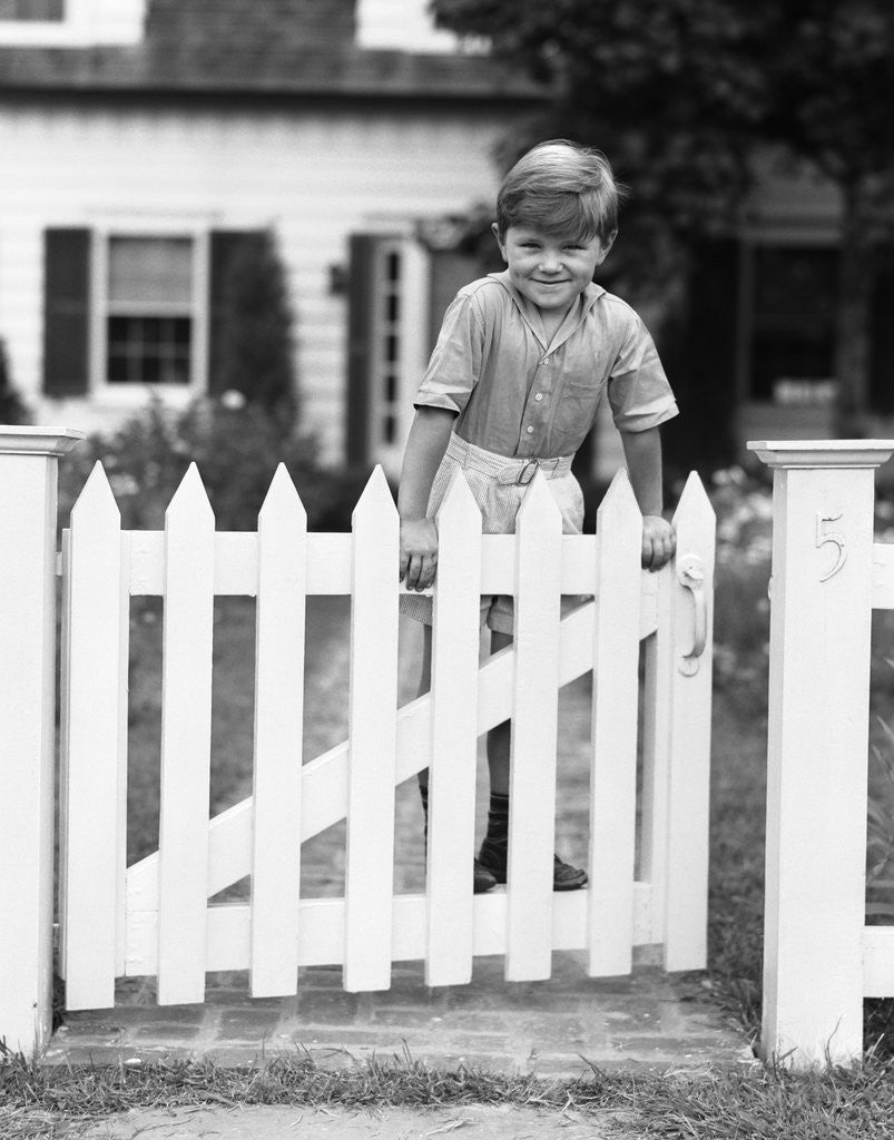 Detail of 1940s Child Boy Standing Swinging On White Picket Fence Gate Smiling Looking At Camera by Anonymous
