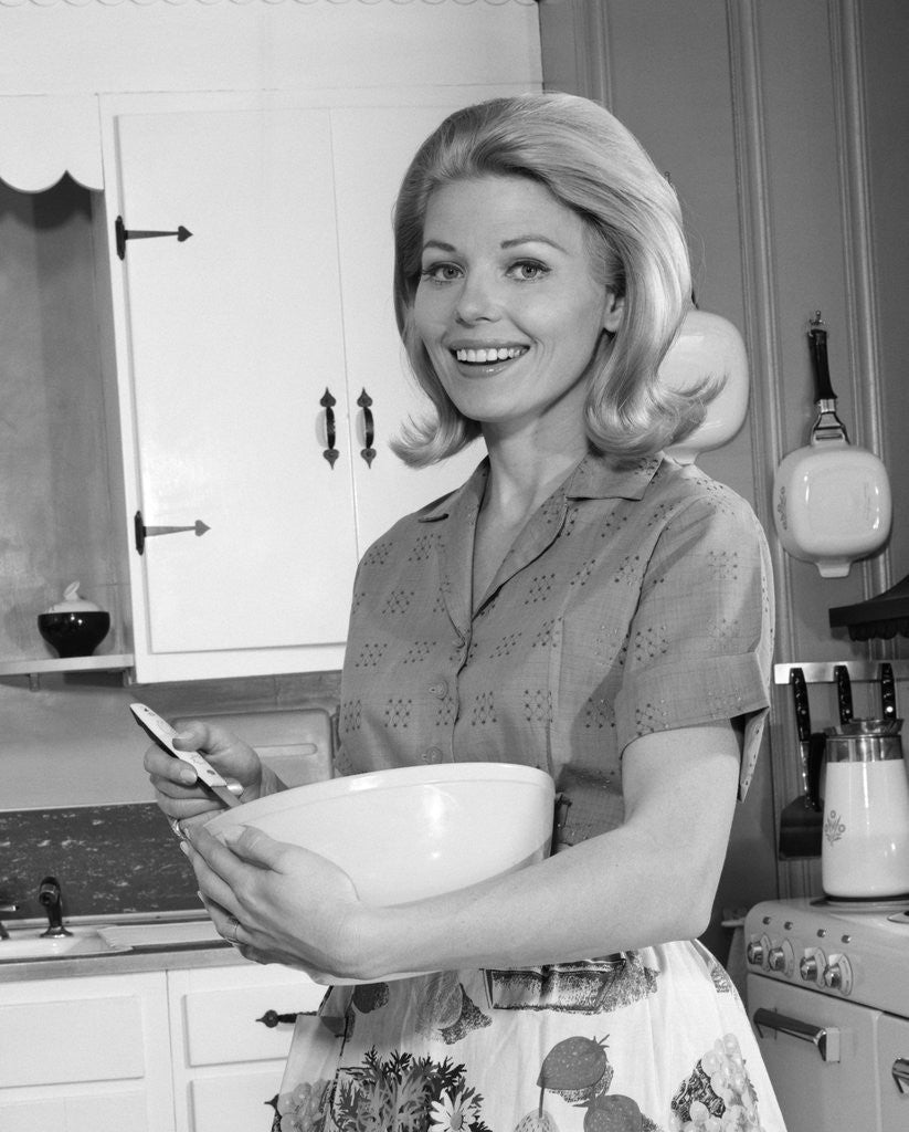 Detail of 1960s 1970s smiling blond housewife in kitchen holding mixing bowl by Anonymous
