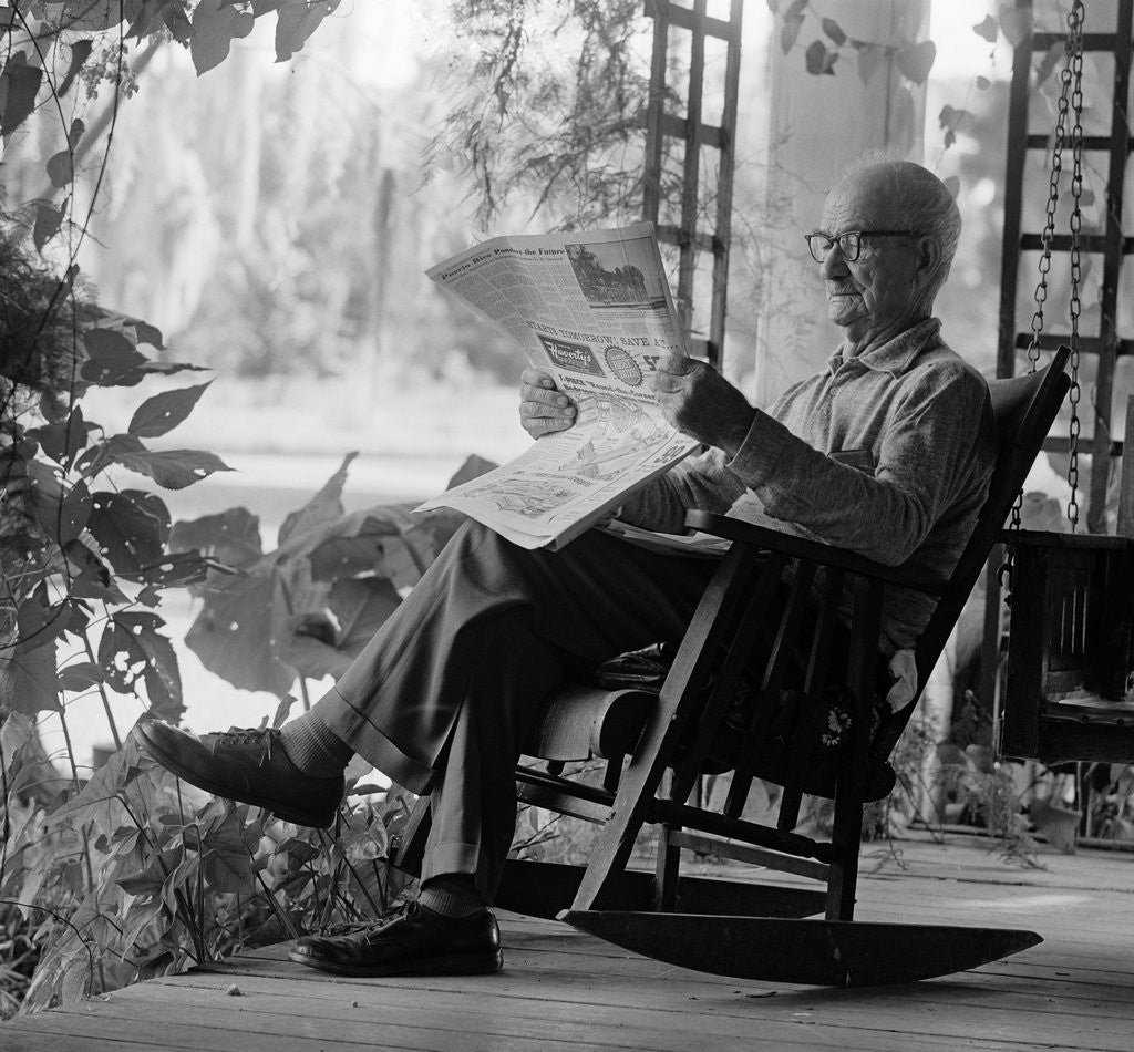 Detail of 1970s elderly man reading newspaper on porch in a rocker by Anonymous