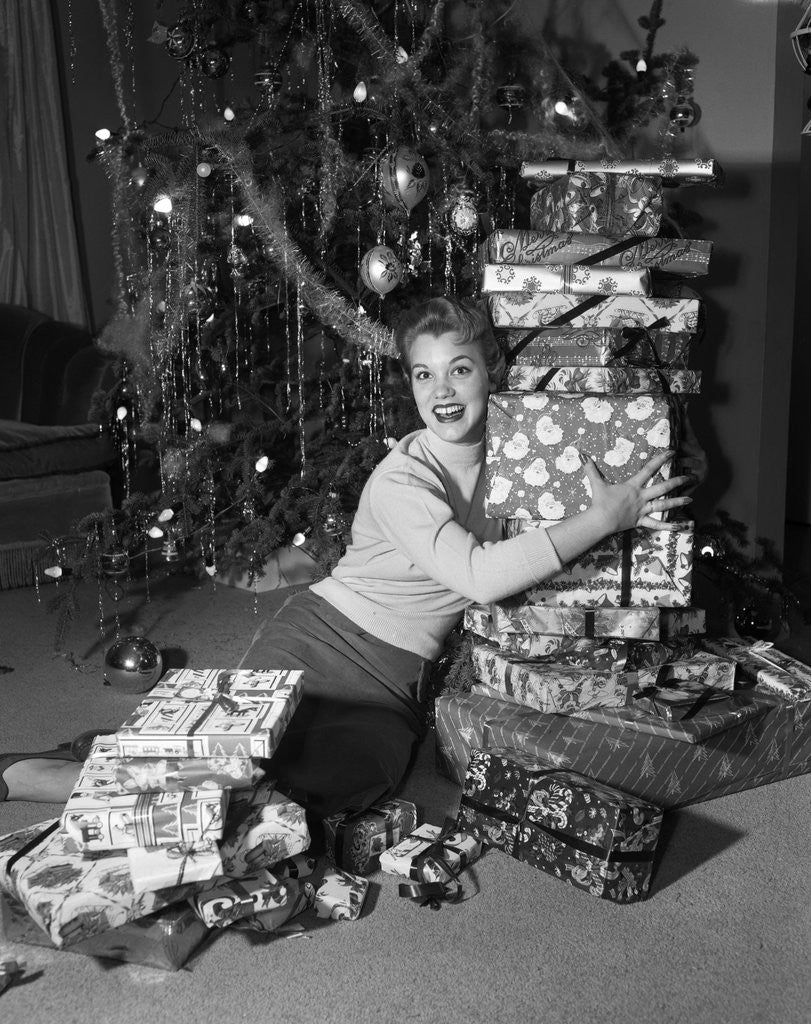 Detail of 1950s portrait of young woman holding stack of presents next to a Christmas tree by Anonymous
